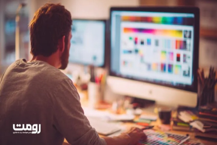 «طراح وب در حال کار روی طراحی رابط کاربری در کامپیوتر رومیزی، محیط کاری مدرن با مانیتورهای بزرگ و نور گرم، لوگوی رومت در گوشه تصویر»